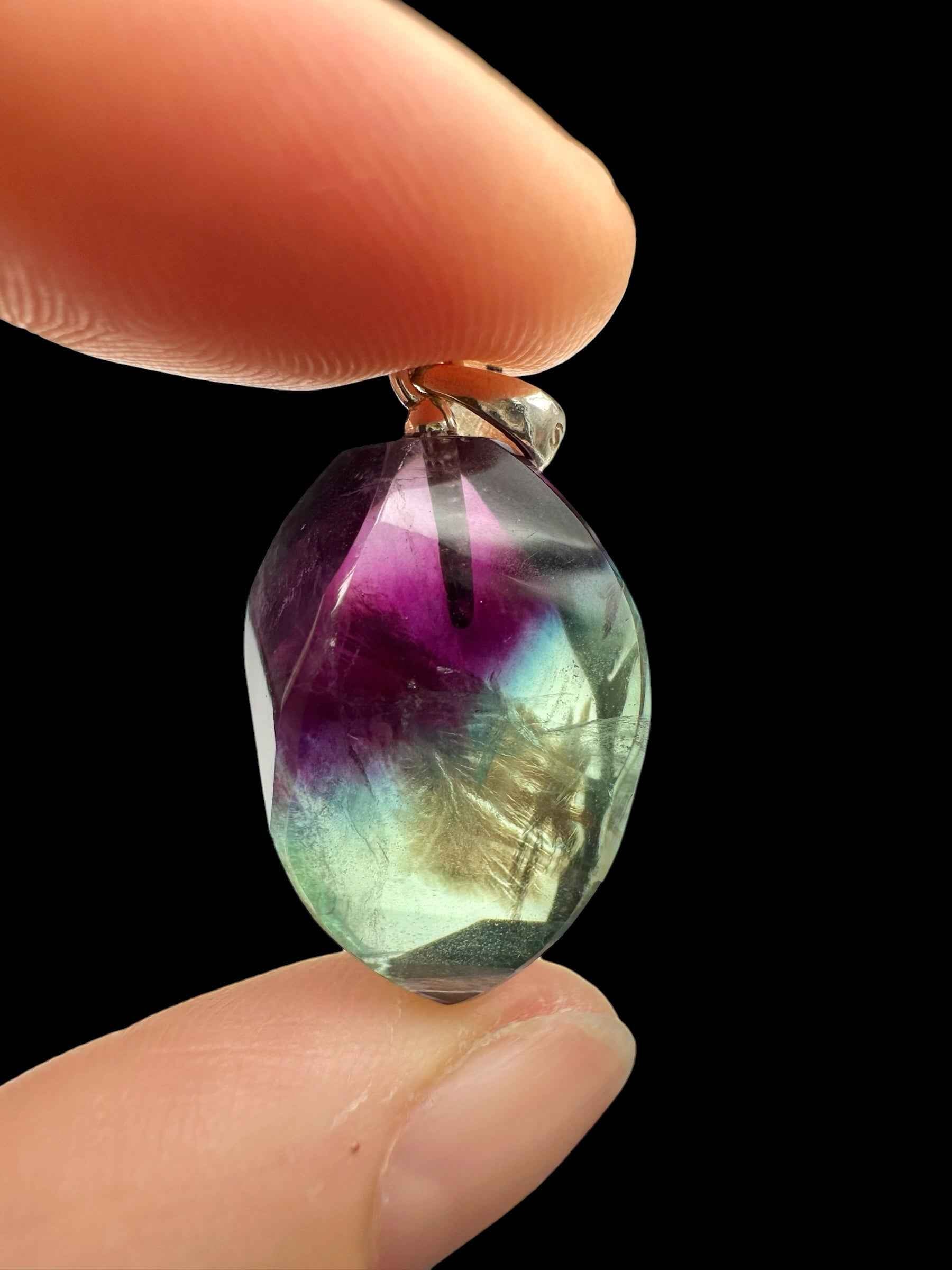 Pendente in fluorite faccettata con zone viola, verde e dorate e archetto in argento, tenuto tra due dita